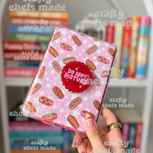 May include: A pink phone case with a pattern of hot dogs and white stars. A red pop socket with the words "DO NOT DISTURB" is attached. The case is held in front of a bookshelf.