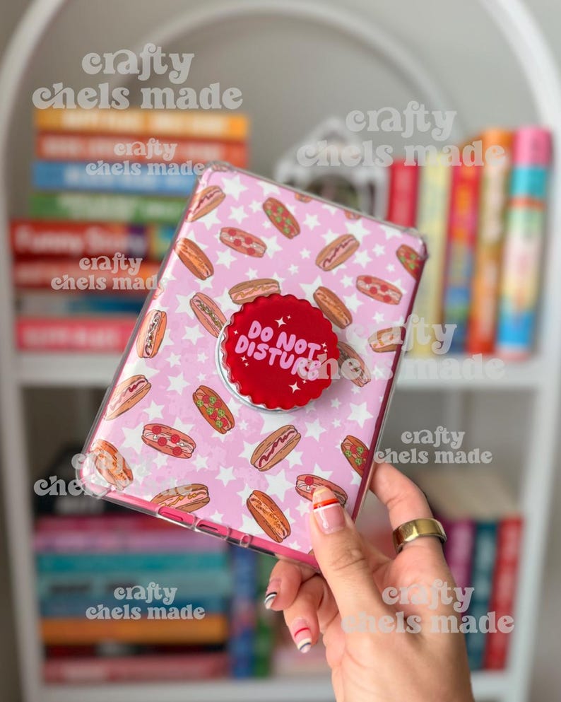 May include: A pink phone case with a pattern of hot dogs and white stars. A red pop socket with the words "DO NOT DISTURB" is attached. The case is held in front of a bookshelf.