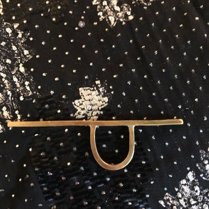 May include: A gold-coloured metal ring with a simple, minimalist design. The ring features a straight bar with a rounded loop at the centre.