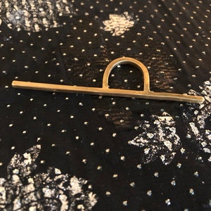 May include: A gold-toned metal ring with a unique design featuring a horizontal bar and a curved arch. The ring is photographed on a black fabric background with a white polka dot pattern.