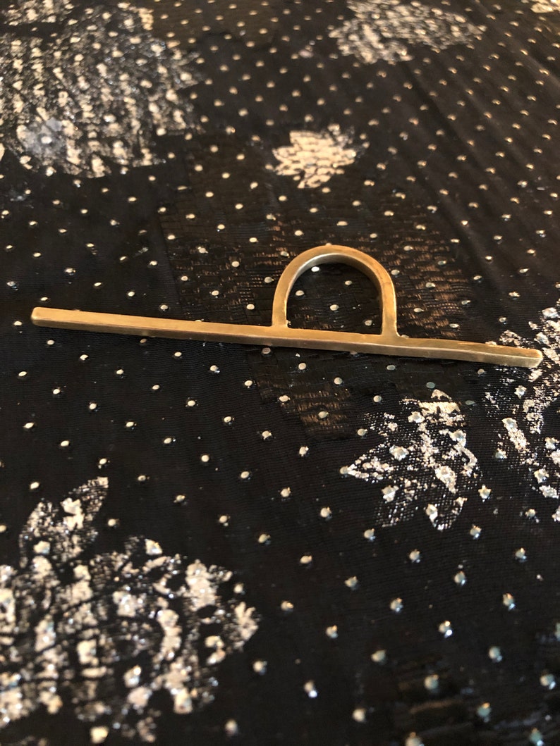 May include: A gold-toned metal ring with a unique design featuring a horizontal bar and a curved arch. The ring is photographed on a black fabric background with a white polka dot pattern.