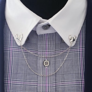 Pode incluir: Close-up de uma camisa branca com gola com presilhas de colarinho prateadas e uma corrente. A camisa tem um padrão xadrez roxo e preto, usada sob um blazer azul marinho. As presilhas de colarinho apresentam um design decorativo.