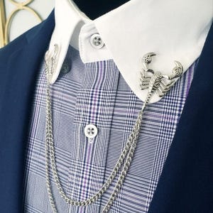 May include: A silver-toned metal collar pin set featuring two scorpion-shaped brooches connected by a double-layered curb chain. The pins are attached to the points of a white shirt collar, which is worn under a blue plaid shirt and a navy blue blazer.