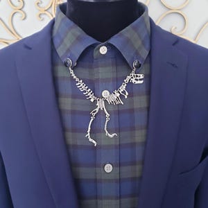 Pode incluir: Uma corrente de colarinho de esqueleto de dinossauro prateado é exibida em uma camisa xadrez azul e verde, usada sob um blazer azul marinho. A corrente do colarinho é presa à gola da camisa com pequenos detalhes pretos.