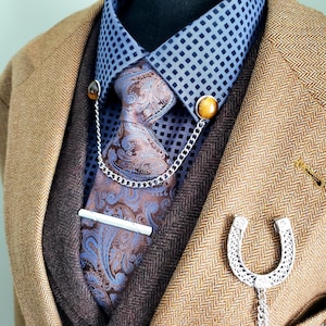 Może przedstawiać: Brązowa marynarka w kratę na ciemnobrązowej kamizelce i niebieskiej koszuli we wzory. Widoczny jest krawat w paisley z srebrnym klipsem do krawata i łańcuszkiem do krawata. Broszka w kształcie podkowy z łańcuszkiem wisi na marynarce.