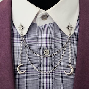 May include: A silver-toned collar pin set featuring two star-shaped brooches with clear crystals. Each brooch is connected by two draped silver chains that hang down to two silver crescent moon charms, attached to a white collared shirt under a purple blazer.