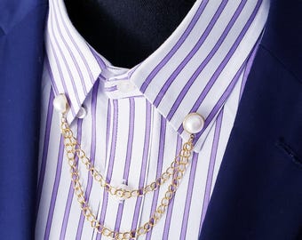 May include: A gold-toned collar chain with faux pearl accents. The chain connects two pearl-topped pins, designed to be worn on a collared shirt. The shirt has a white and purple striped pattern, and is paired with a navy blue blazer.
