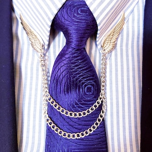 Op de afbeelding: Een close-up van een formele outfit met een paarse stropdas met patroon, een zilveren ketting en vleugelvormige accenten. De stropdas wordt gecombineerd met een gestreept blauw-wit overhemd en een marineblauwe blazer.