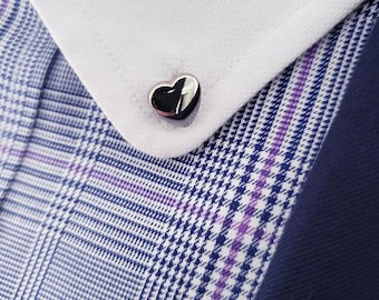 May include: A close-up of a silver heart-shaped cufflink on a white dress shirt cuff. The cuff is set against a blue and purple plaid patterned blazer. The cufflink is shiny and reflective.