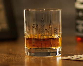 Può includere: Un bicchiere di vetro trasparente con un design di città di Cleveland, Ohio. Il bicchiere è pieno di liquido ambrato e il testo "CLEVELAND" è stampato sul fondo.