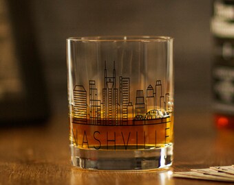 May include: A clear glass tumbler with a black outline of the Nashville skyline and the word "NASHVILLE" printed on the bottom. The glass is filled with a brown liquid.
