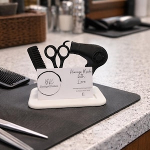 May include: A white rectangular holder with black hair styling tools, including scissors, a comb, and a hairdryer. A business card with the text "Always Made with Love" is also in the holder. The holder sits on a black mat.