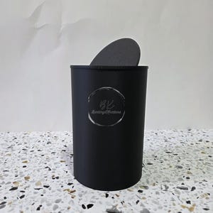 May include: A black cylindrical container with a dark gray lid. The container has a silver label with the text "BK" and "Barlings Kreations". The container is on a speckled surface.