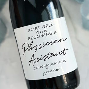 May include: A white champagne bottle label with black text that reads "Pairs well with becoming a Physician Assistant. Congratulations Jenna".
