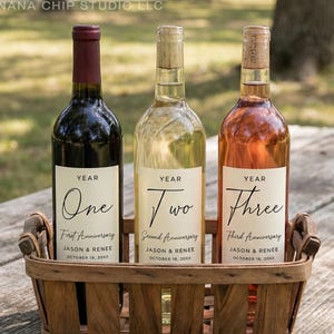 May include: Three wine bottles in a wooden basket. The bottles are labeled "Year One", "Year Two", and "Year Three" with anniversary details. The bottles are filled with red, white, and rosé wine. The basket is on a wooden picnic table.