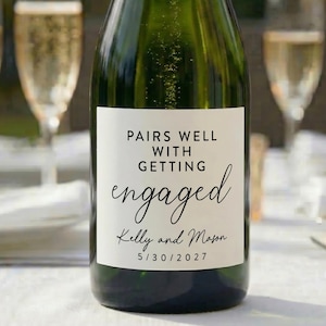 May include: A green champagne bottle with a gold foil top and a white label. The label reads "Pairs Well With Getting Engaged" with the names "Kelly and Mason" and the date "5/30/2027".