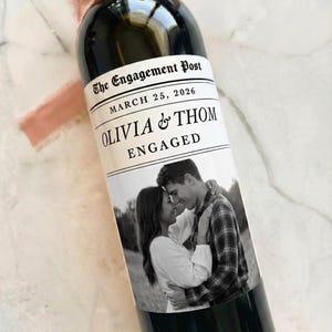 May include: A dark wine bottle with a custom label that reads "OLIVIA & THOM ENGAGED" with a photo of a couple. The label also includes the date March 25, 2026, and the text "The Engagement Post". A pink ribbon is tied around the bottle's neck.