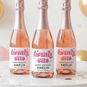 May include: Three glass bottles filled with a rose-colored liquid, each featuring a white label with the pink text twenty one, the words HAPPY BIRTHDAY AMELIA, and the date 02.02.20XX. The bottles have metallic rose gold foil neck wraps.