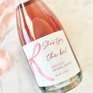 May include: A glass bottle filled with a rose-colored liquid, featuring a white label with a pink ribbon illustration and the text "She's tying the knot", "HOLLIE'S BRIDAL SHOWER", and "MAY 2024". A frayed, light pink silk ribbon is tied around the bottle neck.