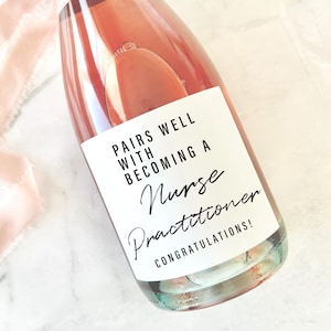 May include: A champagne bottle with a pink label that reads "PAIRS WELL WITH BECOMING A Nurse Practitioner CONGRATULATIONS!". The bottle contains a pink liquid and is decorated with a pink ribbon. The bottle size is approximately 10.16cm x 8.26cm.