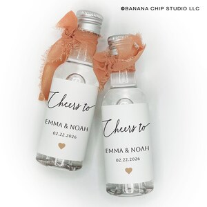 May include: Two clear glass mini bottles with silver screw caps, each tied with a frayed peach-colored ribbon. The white labels feature the black text Cheers to EMMA & NOAH 02.22.2026 and a small gold heart icon. The text BANANA CHIP STUDIO LLC is visible.