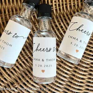May include: Three clear glass miniature bottles with black screw caps, each featuring a white label printed with the text Cheers to EMMA & THOM 5.20.2025 and a small peach-colored heart icon. The bottles are empty and have a narrow neck design.