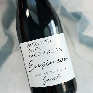 May include: A champagne bottle label with the text "Pairs well with becoming an Engineer. Congratulations Jacob." The label is white with black text and is on a dark green bottle.