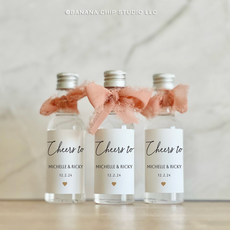 May include: Three clear glass miniature bottles with silver screw caps, each adorned with a frayed peach-colored ribbon. The white labels feature the black text Cheers to, MICHELLE & RICKY, the date 12.2.24, and a small gold heart icon.