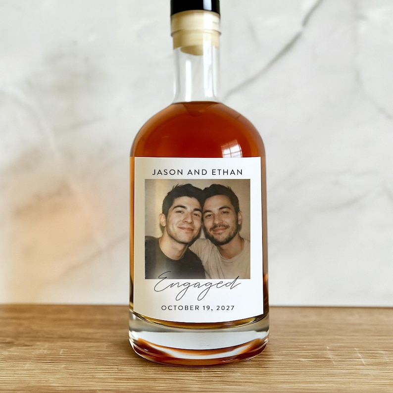 May include: A clear glass bottle filled with amber liquid, featuring a label with a photo of two men. The label reads "Jason and Ethan," with the word "Engaged" and the date "October 19, 2027" below the photo.