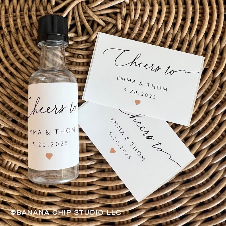 May include: A clear plastic bottle with a black cap and two rectangular white labels. The labels feature the black text Cheers to EMMA & THOM 5.20.2025 and a small peach-colored heart. The bottle and labels are displayed together as a set.