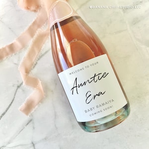 May include: A white label on a bottle of pink champagne with the text "Welcome to your Auntie Era Baby Ramaiya Coming Soon!"