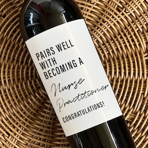 May include: A bottle of red wine with a white label. The label reads "PAIRS WELL WITH BECOMING A Nurse Practitioner CONGRATULATIONS!" The bottle is set on a woven, circular, brown surface.