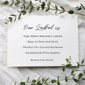 May include: A white card with the text "Our Label is" in elegant script. Below, bullet points list features: high water resistance, easy peel and stick, perfect for curved surfaces, and exceptional print quality. The card is surrounded by greenery.