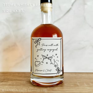 May include: A clear glass whiskey bottle with a label that reads "Pairs well with getting engaged." The label includes illustrations of champagne glasses, a heart-shaped box, and a champagne bottle. The bottle is 4 inches by 3.5 inches.