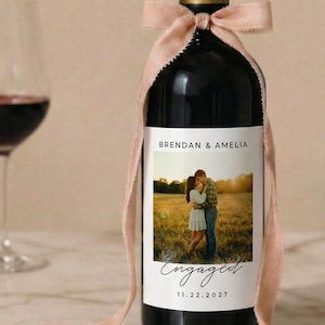 May include: A dark wine bottle with a custom label featuring a photo of a couple and the word "Engaged." The label also includes the names "Brendan & Amelia" and the date "11.22.2027." A blush pink ribbon is tied around the neck of the bottle.