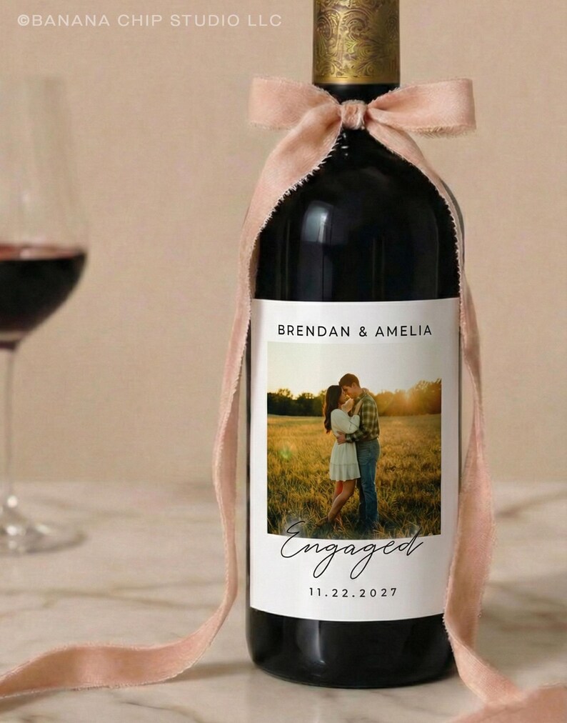 May include: A dark wine bottle with a custom label featuring a photo of a couple and the word "Engaged." The label also includes the names "Brendan & Amelia" and the date "11.22.2027." A blush pink ribbon is tied around the neck of the bottle.