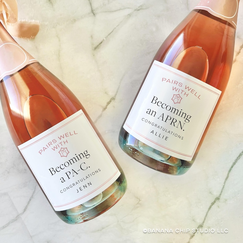 May include: Two bottles of sparkling wine with pink labels that read "Pairs Well With" and "Becoming a PA-C. Congratulations Jenn" and "Becoming an APRN. Congratulations Allie". The bottles are on a marble surface.