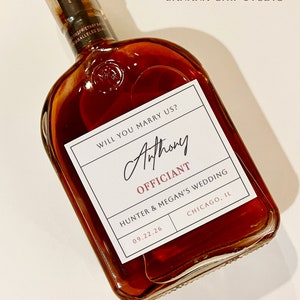 May include: A square-shaped glass bottle filled with amber liquid, featuring a white label with the text "WILL YOU MARRY US?" and the name "Anthony" in elegant script. The label also includes "OFFICIANT", wedding details, and the date 09.22.26.