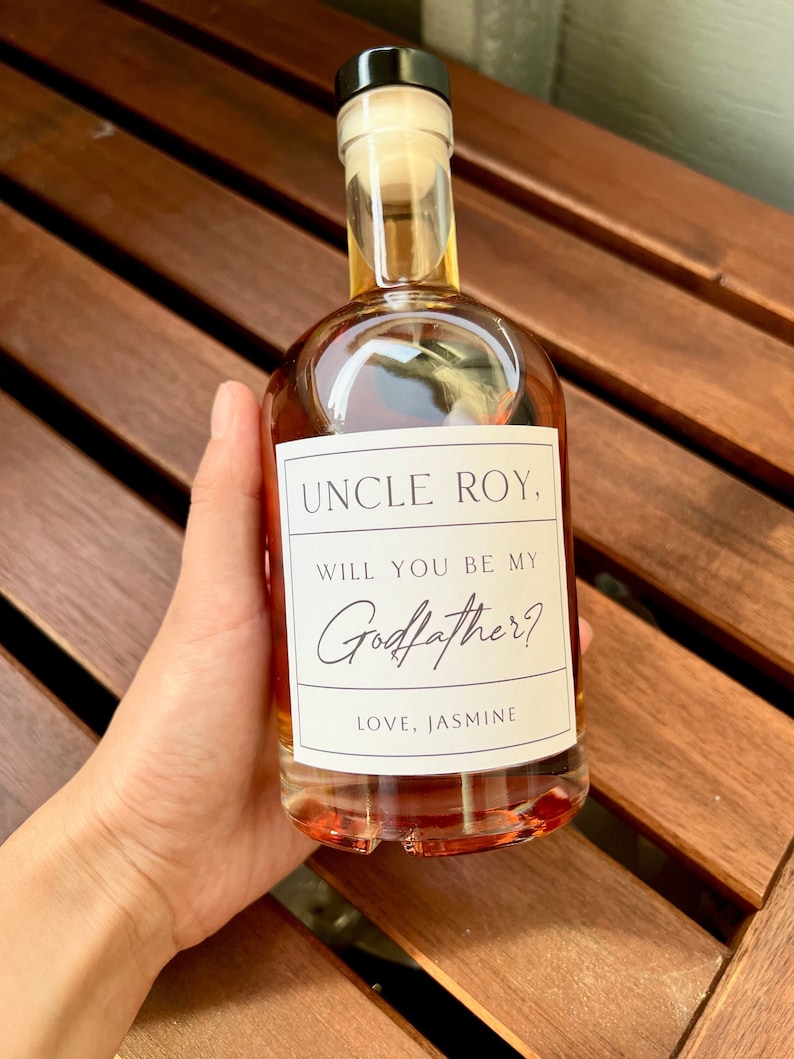 May include: A clear glass bottle with an amber liquid, a black cap, and a white label. The label says "UNCLE ROY, WILL YOU BE MY Godfather? LOVE, JASMINE". The bottle is held against a wooden surface, ideal for a gift.