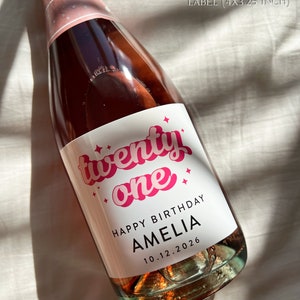 May include: A 750ml champagne bottle with a custom white label featuring the pink text 'twenty one', 'HAPPY BIRTHDAY AMELIA', and the date '10.12.2026'. The label measures 4x3.25 inches and includes small pink star icons around the text.