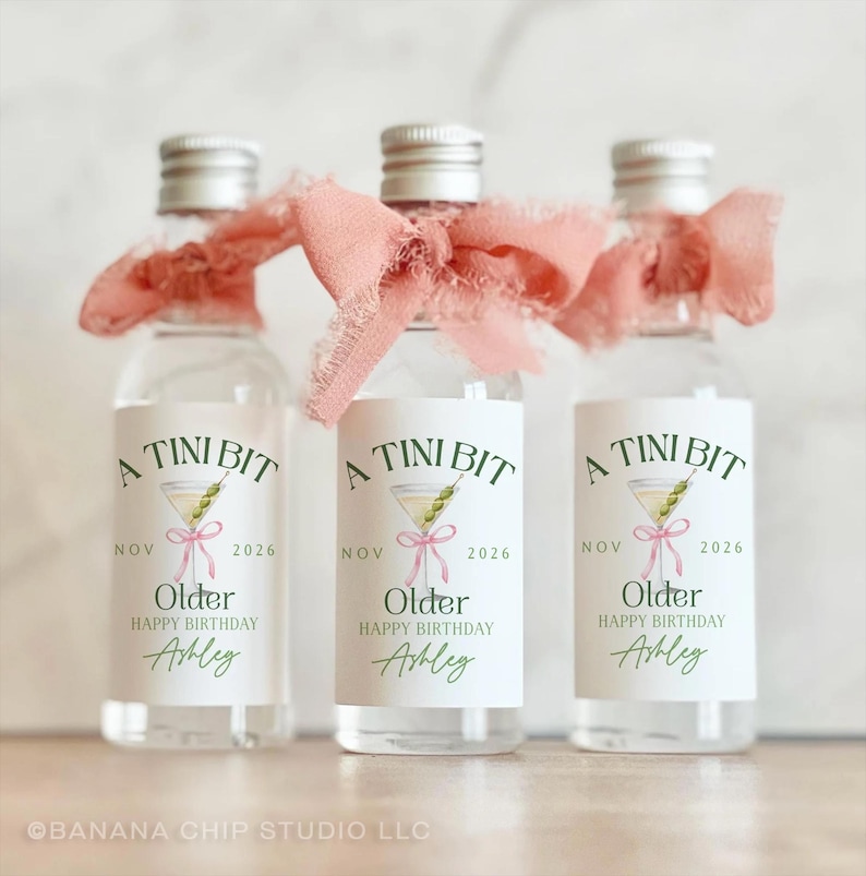 Puede incluir: Tres peque&ntilde;as botellas de vidrio con tapas plateadas, cada una adornada con una cinta rosa. Las etiquetas dicen "A Tini Bit" con una ilustraci&oacute;n de martini, "Older Happy Birthday Ashley" y la fecha "Nov 2026".