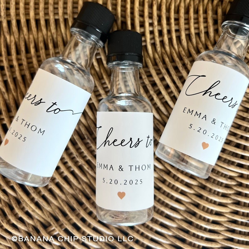 May include: Three clear glass miniature bottles with black screw caps, each featuring a white label printed with the text Cheers to EMMA & THOM 5.20.2025 and a small peach-colored heart icon. The bottles are empty and have a narrow neck design.