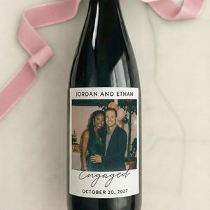 May include: A black wine bottle with a custom label featuring a photo of a couple. The label reads "JORDAN AND ETHAN" above the photo and "Engaged" with the date "OCTOBER 20, 2027" below. A pink ribbon is tied around the bottle's neck.