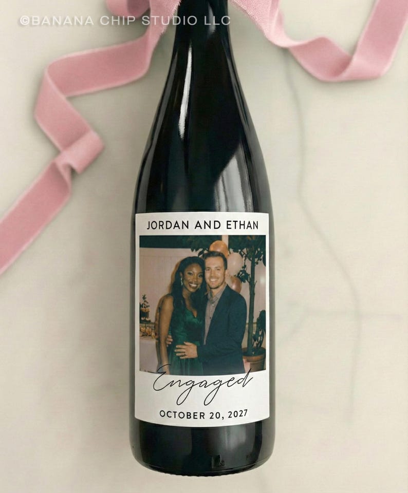 May include: A black wine bottle with a custom label featuring a photo of a couple. The label reads "JORDAN AND ETHAN" above the photo and "Engaged" with the date "OCTOBER 20, 2027" below. A pink ribbon is tied around the bottle's neck.