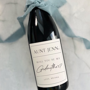 May include: A black wine bottle with a white label that reads "AUNT JENN, WILL YOU BE MY Godmother? LOVE, BRIANA." The bottle is tied with a light blue ribbon. The bottle is 8.89 x 10.16 cm and holds 750ml of liquid.