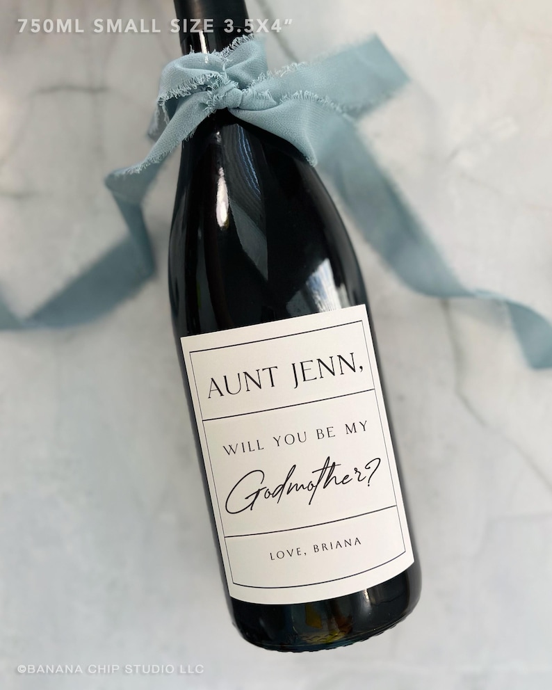 May include: A black wine bottle with a white label that reads "AUNT JENN, WILL YOU BE MY Godmother? LOVE, BRIANA." The bottle is tied with a light blue ribbon. The bottle is 8.89 x 10.16 cm and holds 750ml of liquid.