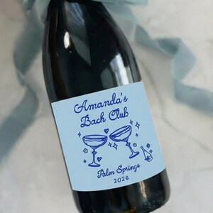 May include: A dark glass champagne bottle with a light blue label featuring the text Amanda's Bach Club, Palm Springs 2026, and illustrations of two champagne coupes and a bottle. The label measures 4x3.25 inches and a blue ribbon is tied at the neck.