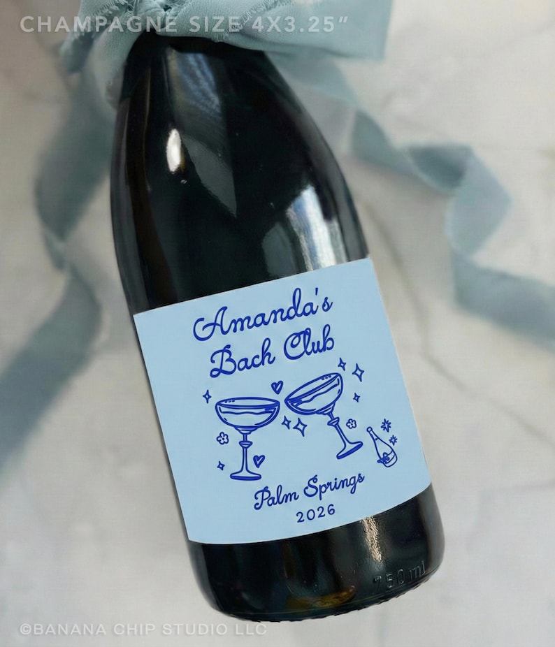 May include: A dark glass champagne bottle with a light blue label featuring the text Amanda's Bach Club, Palm Springs 2026, and illustrations of two champagne coupes and a bottle. The label measures 4x3.25 inches and a blue ribbon is tied at the neck.