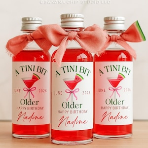 Puede incluir: Tres botellas de vidrio peque&ntilde;as llenas de un l&iacute;quido rojo brillante, cada una con una tapa plateada y una cinta color coral. Las etiquetas dicen "A Tini Bit" con una ilustraci&oacute;n de c&oacute;ctel, "Junio 2026", "Older Happy Birthday Nadine".