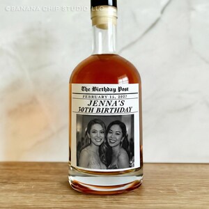 May include: A clear glass bottle filled with a brown liquid, likely a spirit. The label reads "The Birthday Post" with the date "February 15, 2027" and "Jenna's 30th Birthday." The label also features two women in a black and white photo.